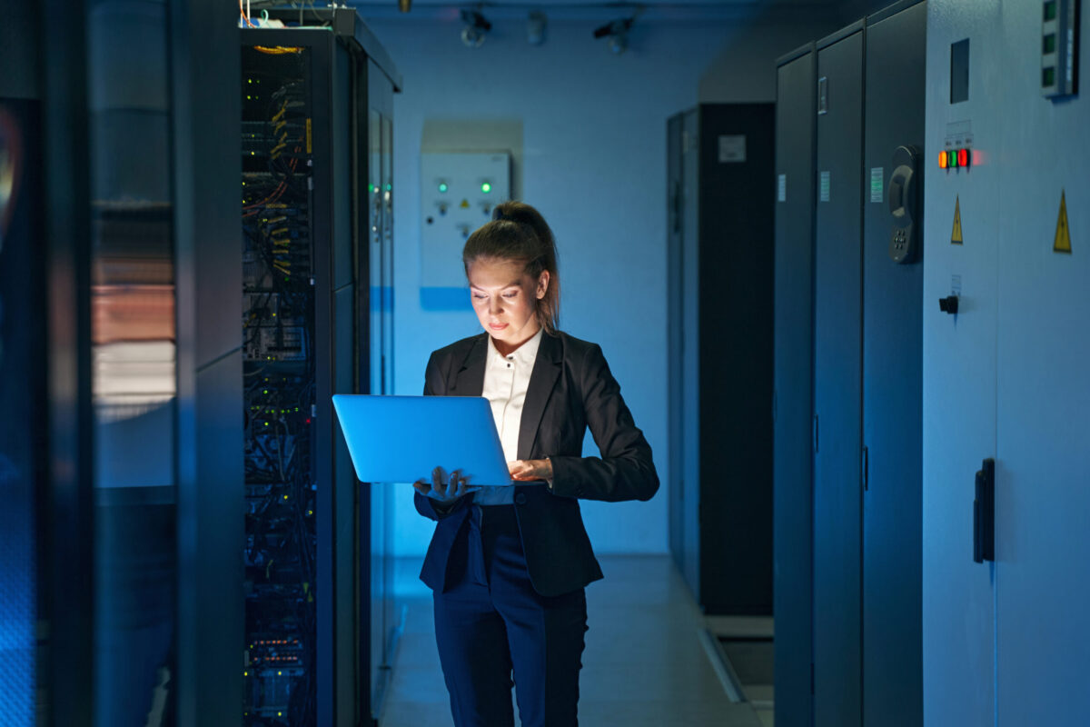 Young woman engineer with laptop computer working at network equipment between server racks in data center