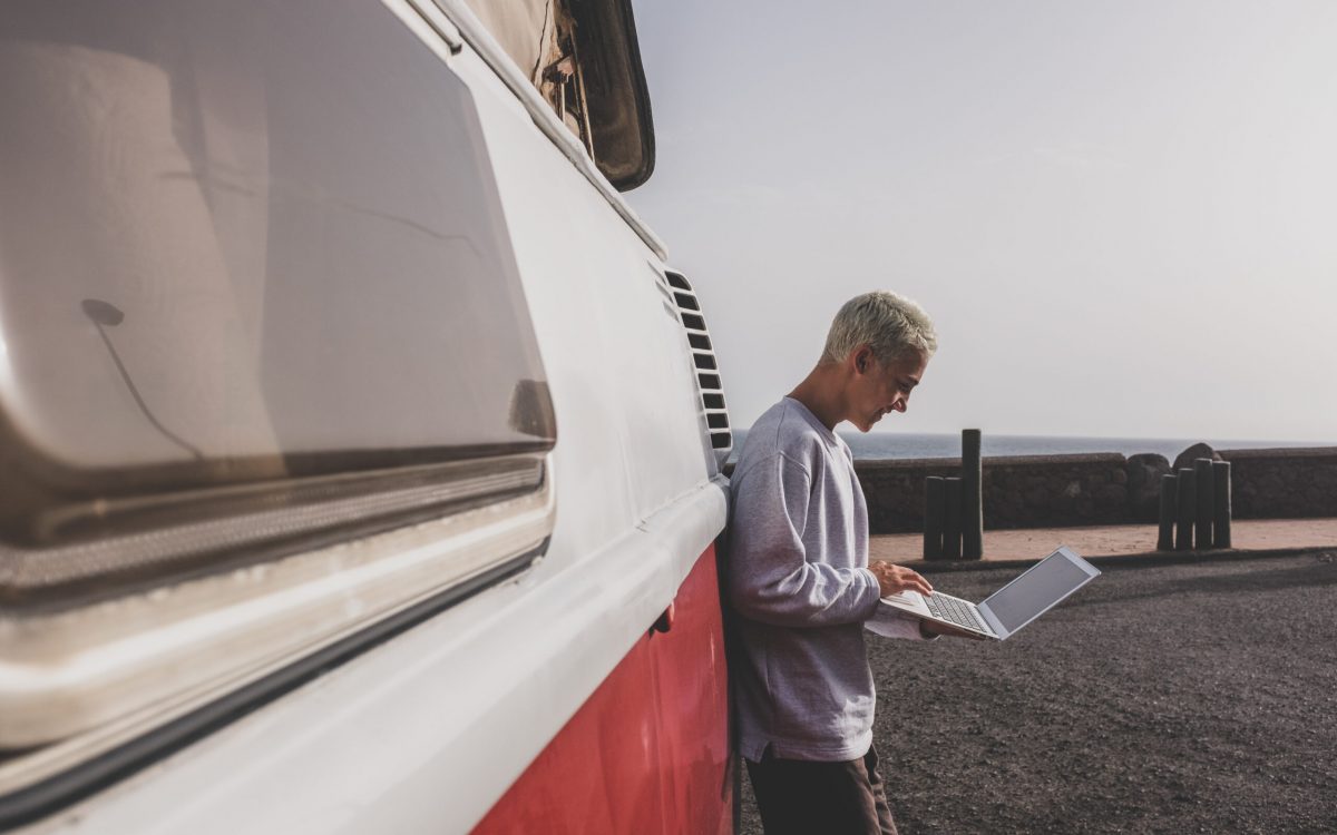 one young man using his laptop or computer pc smiling and looking at it - technology nomad lifestyle traveling and discovering the world with his red mini van and his device to work